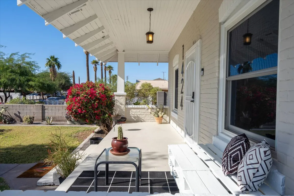 An inviting front porch features comfortable outdoor seating, decorative pillows, and lush flowering plants. The arrangement encourages relaxation or socializing while overlooking the neighborhood, all under abundant natural light.