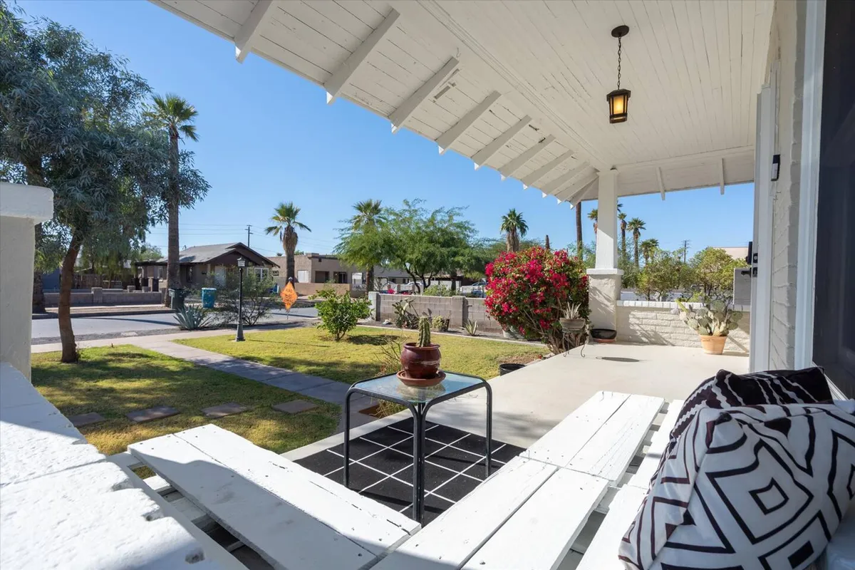 A cheerful exterior view of the front porch with a potted plant, blooming bougainvillea, and comfortable outdoor furniture. The setup beckons for relaxation or casual socializing, blending indoor and outdoor living.