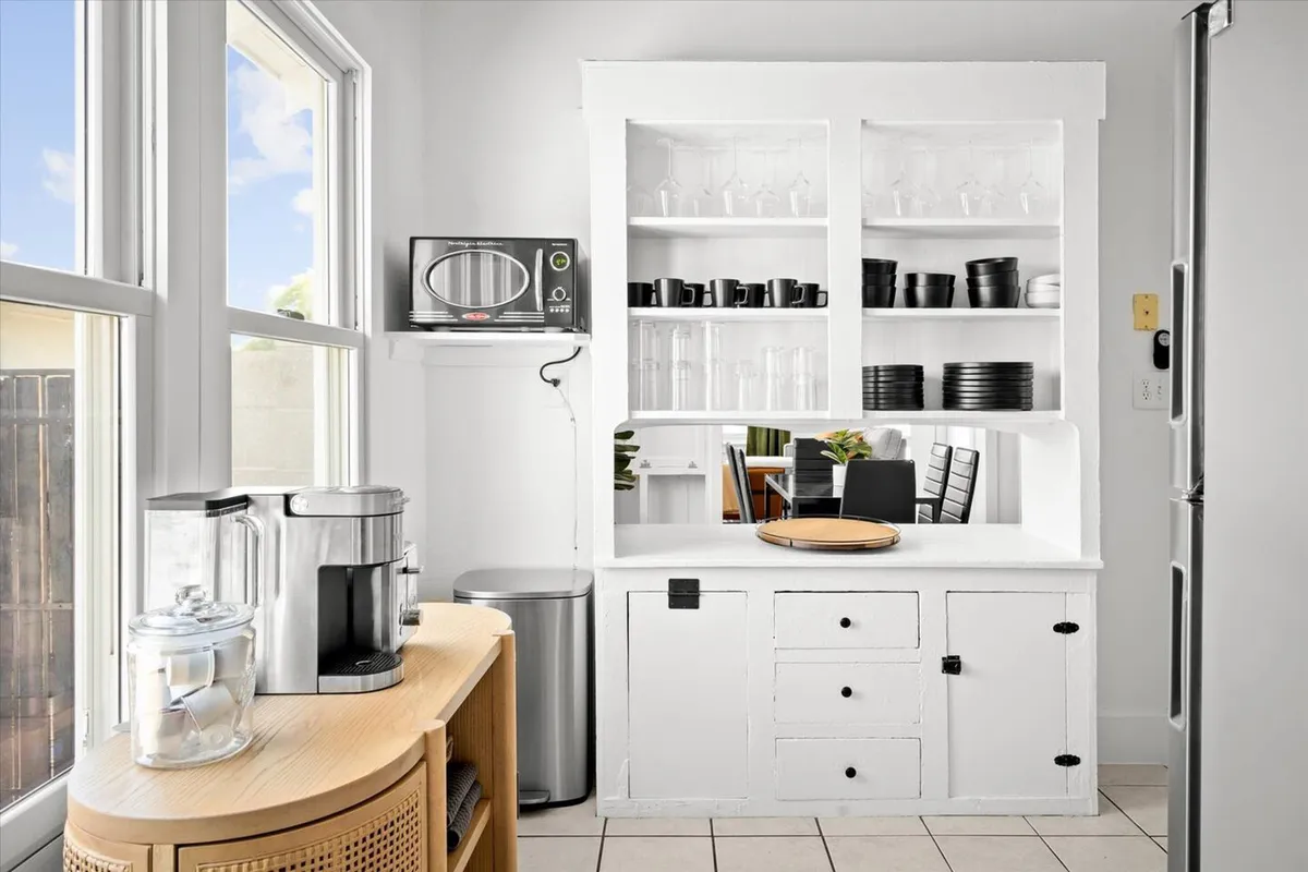 A view of sleek white cabinetry, open shelving with glassware and black dishware, and a coffee maker situated on the counter. The space feels cozy yet functional, highlighting both the stocked cabinets and the ease of making coffee or casual meals.