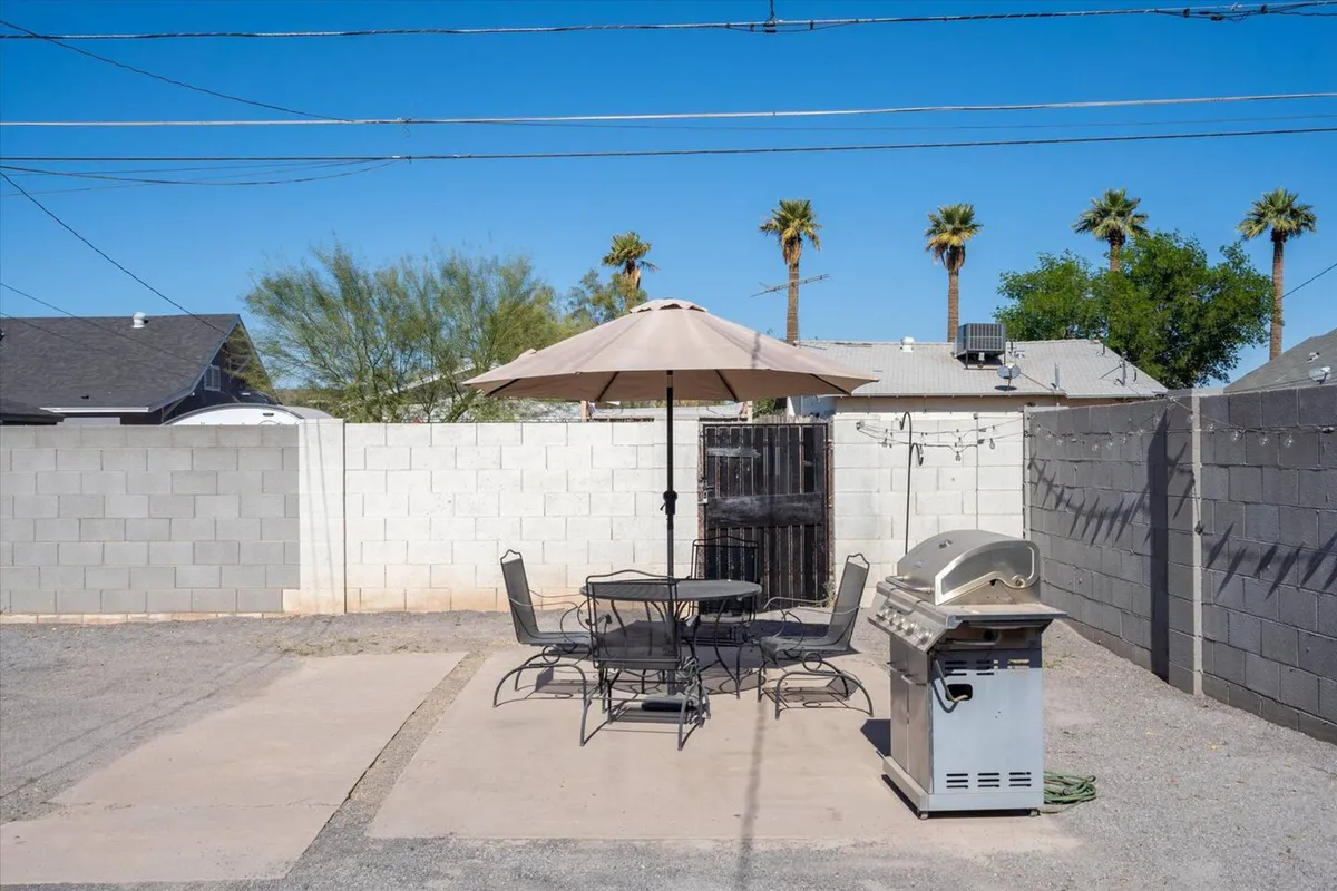 A backyard scene featuring a patio table with an umbrella, a barbecue grill, and palm trees in the background. The area is set for outdoor dining and casual gatherings, evoking a sense of community and leisure.