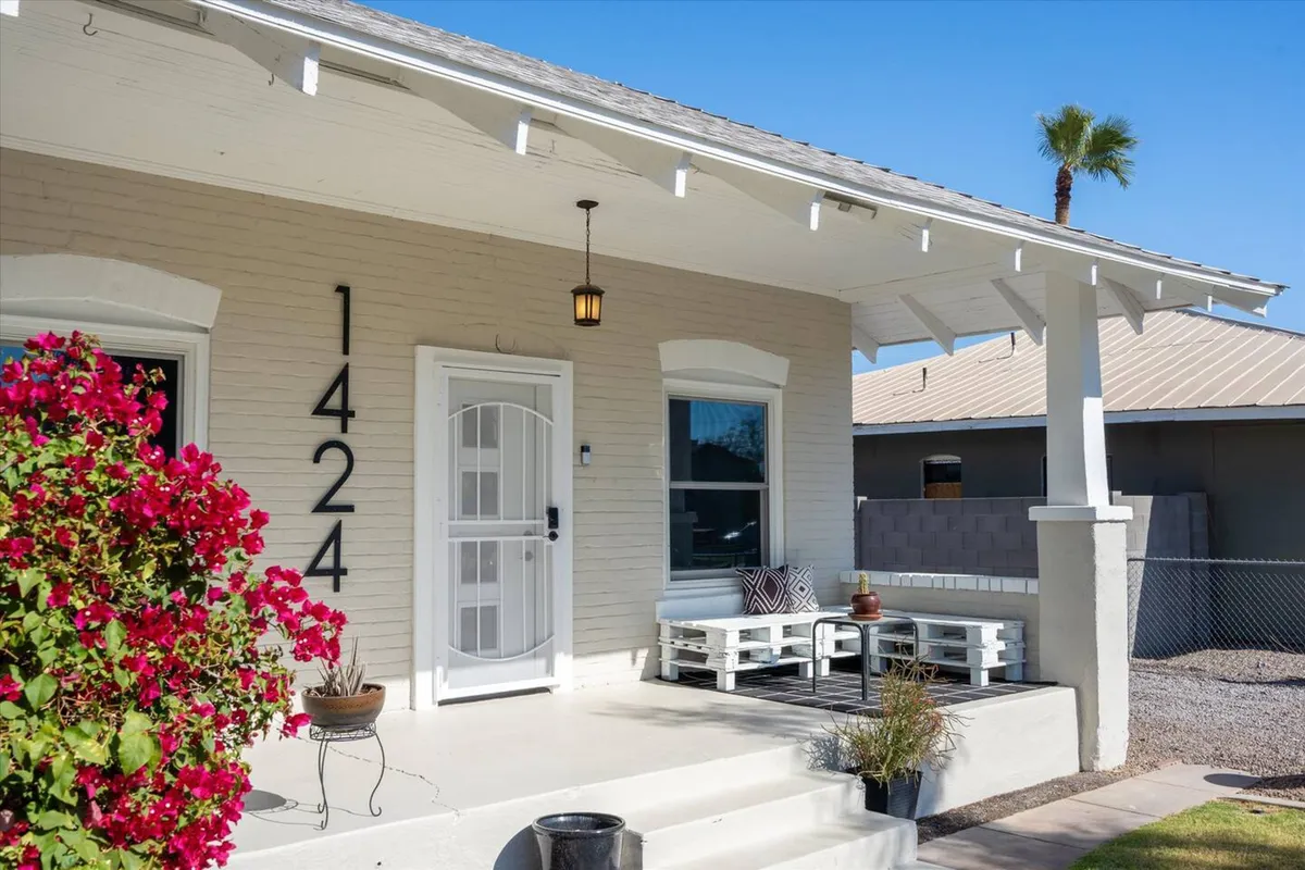 A welcoming exterior shot showing the house number 1424, vibrant potted plants, a flowering bush, and a cozy seating area on the front porch. The scene is bright and inviting, capturing the sense of arrival and neighborhood context.