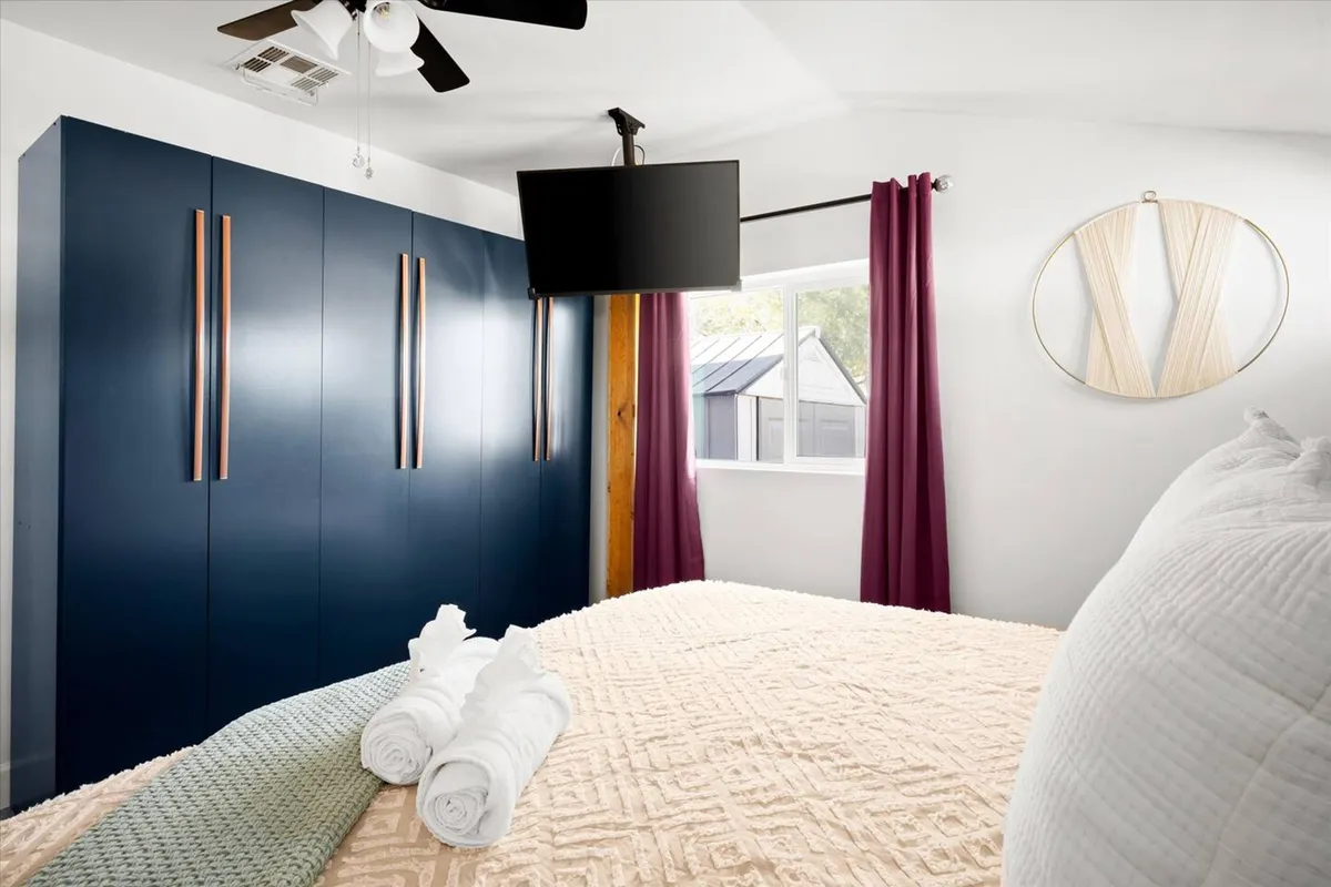 A welcoming bedroom featuring a bed adorned with decorative pillows, a blue wardrobe offering ample storage, a television for unwinding, and soft curtains framing a window. The atmosphere is inviting and comfortable, ideal for relaxation and privacy.