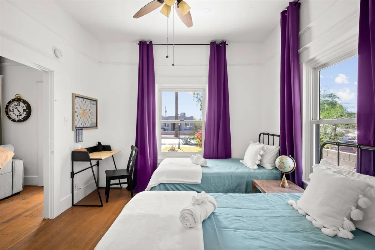 A cheerful bedroom featuring two beds with turquoise bedding, purple curtains, and a world globe on a table. The room is bright and relaxed, suitable for both families and professionals needing flexibility. The window welcomes morning light, enhancing the calm mood.