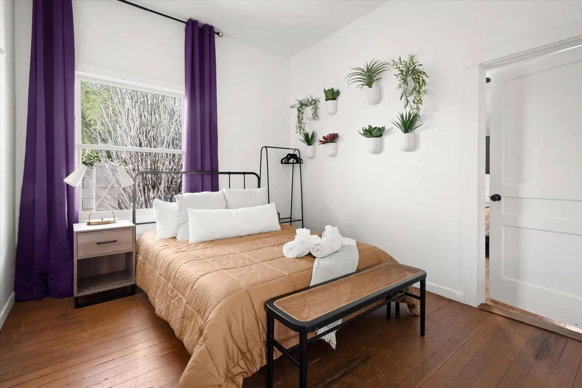 A serene bedroom showcasing a bed with neutral bedding, purple curtains gently filtering natural light, wall-mounted planters adding a touch of greenery, and a nightstand with a lamp for nighttime reading. The overall atmosphere is calm and inviting.