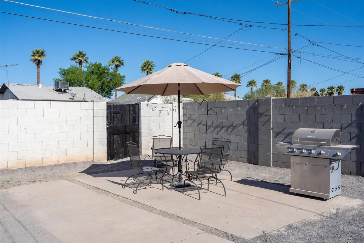 An outdoor scene featuring a table with an umbrella, a grill, and a black gate. The area is set up for dining and social gatherings, with plenty of shade and a welcoming ambiance for family barbecues or relaxed afternoons.
