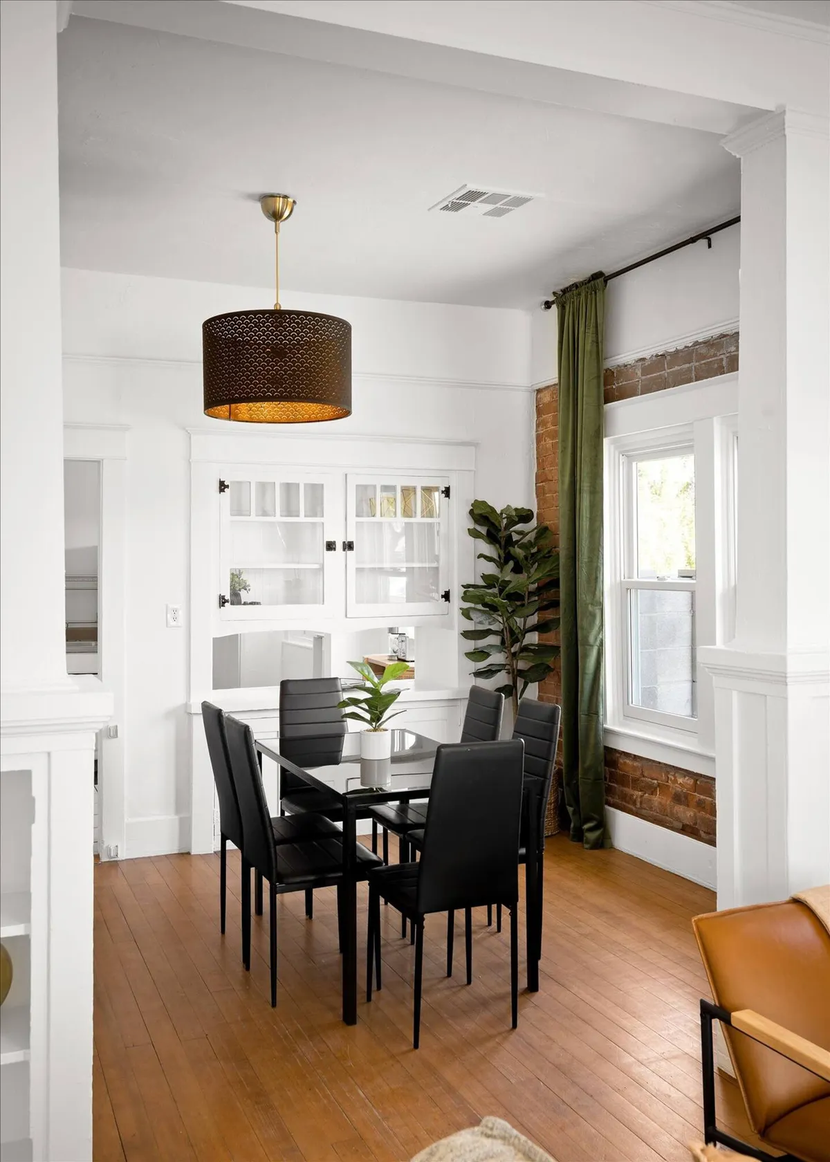 A contemporary dining area featuring a spacious dining table set beneath a striking hanging light fixture, with green curtains and an indoor plant adding warmth. The exposed brick wall adds a touch of modern charm, making the space perfect for family dinners or creative gatherings.